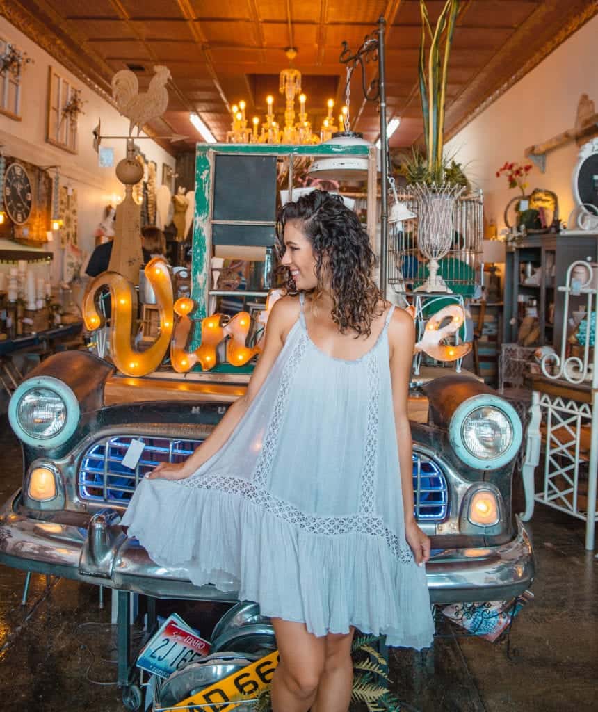 Jessica in a flowy white dress stands by a vintage car
