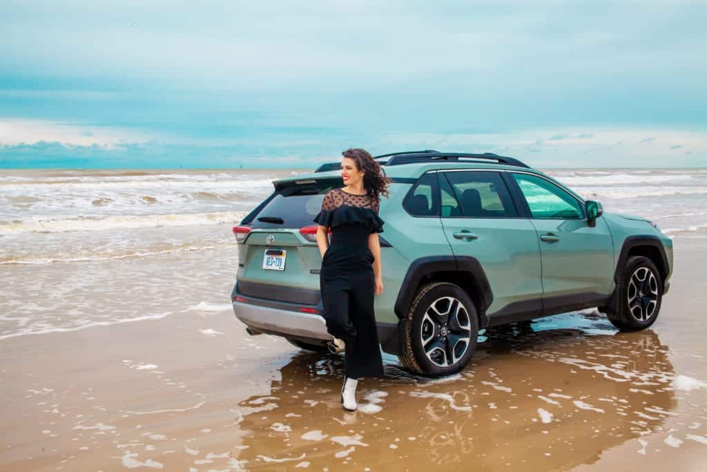 A teal SUV parked on the beach with waves touching the tires.