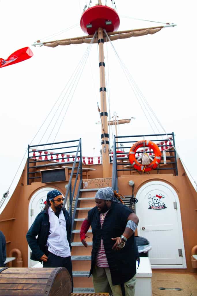 Two people standing on a pirate-themed ship deck with a lifebuoy and a mast.