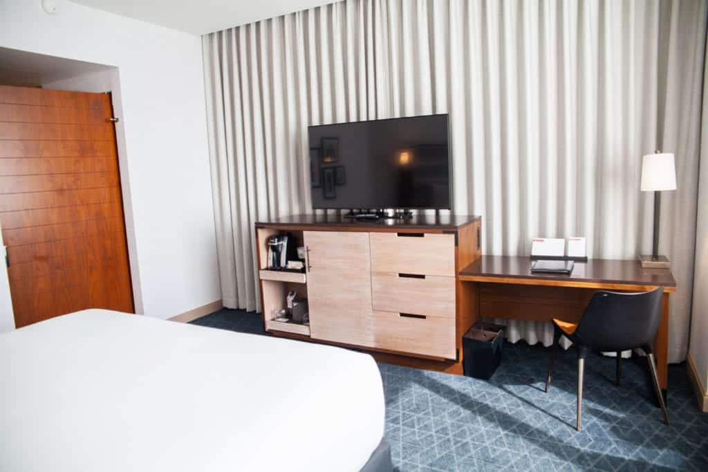 Modern hotel room with a bed, TV, desk, and chair.