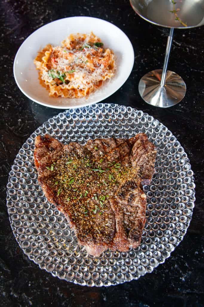 a steak on a plate next to a plate of pasta and a glass of wine