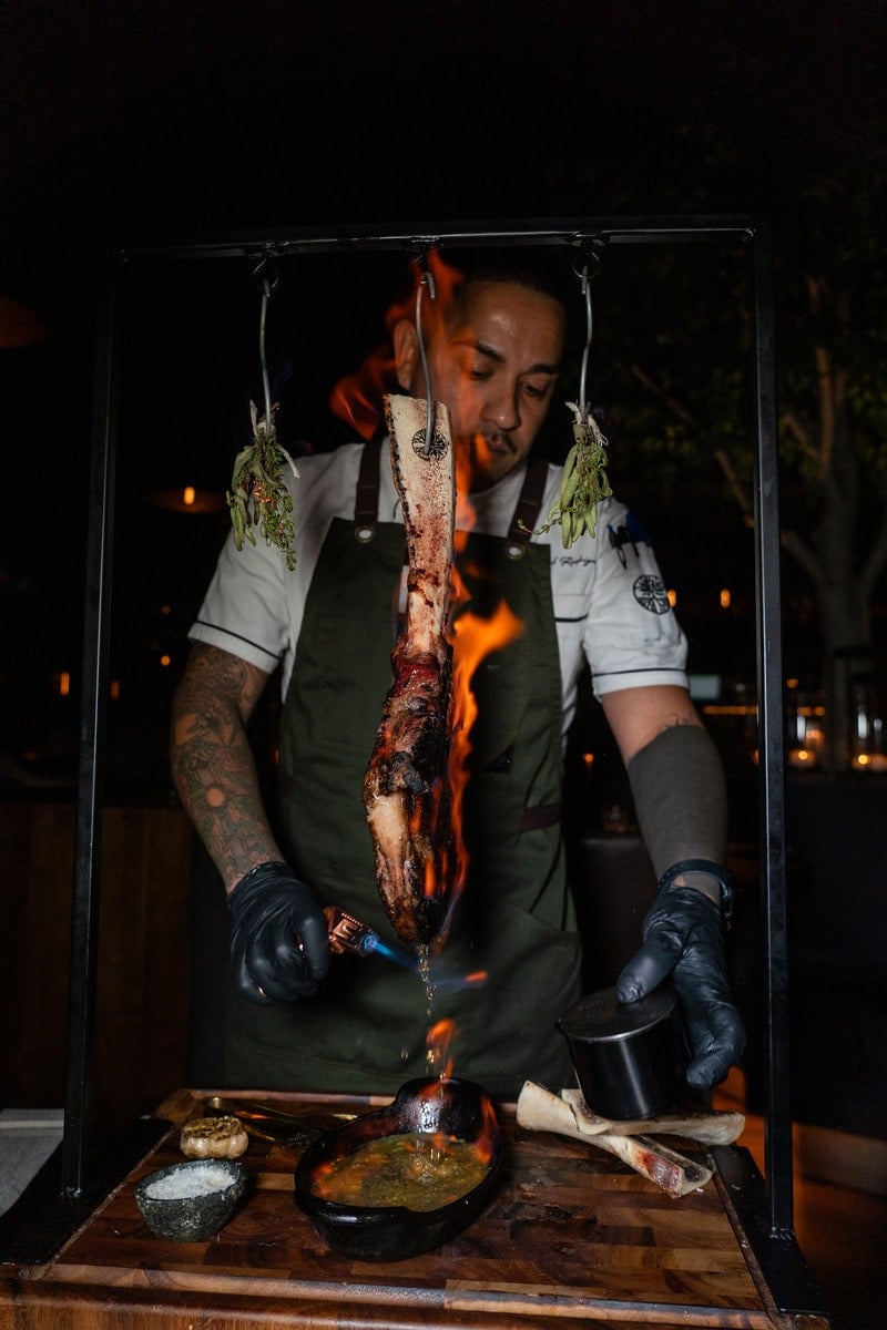A waiter in an apron and black gloves preparing a hanging tomahawk with fire and herbs tableside