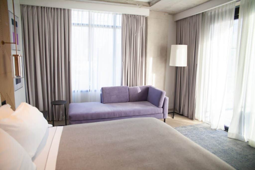 Modern hotel room featuring a gray bed, a purple sofa, and large windows dressed with sheer curtains at canopy hotel