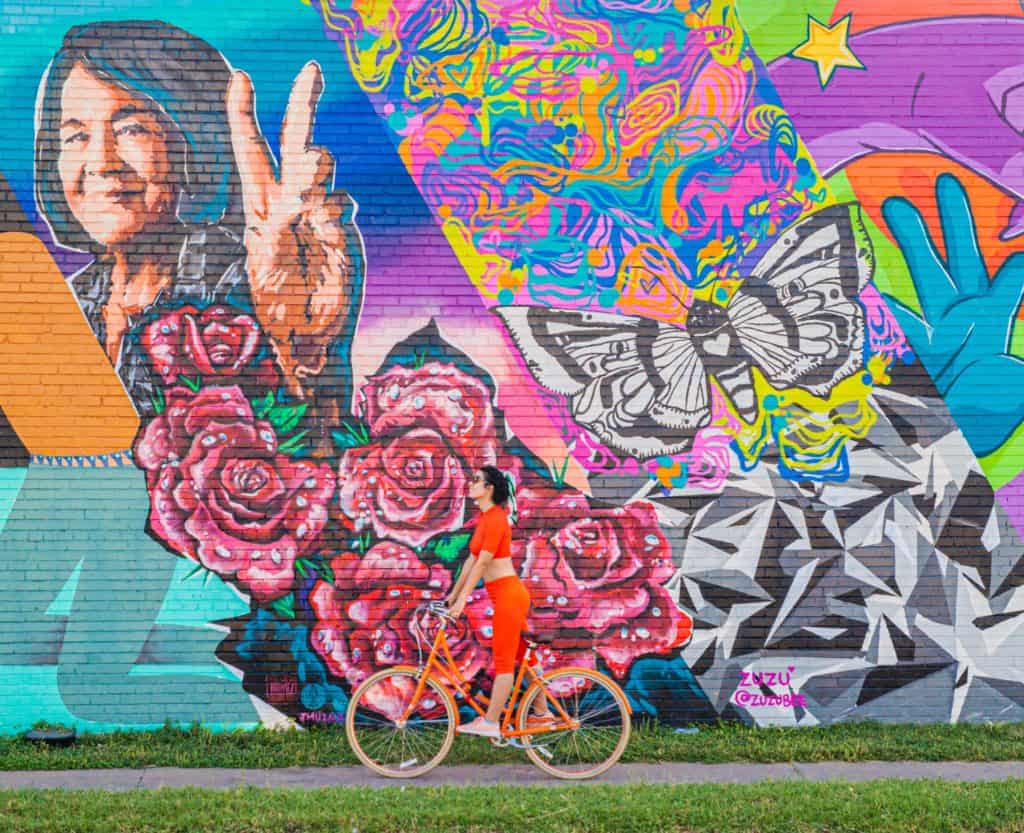 Jessica rides a bicycle past a colorful mural featuring roses, butterflies, and abstract designs near canopy hotel