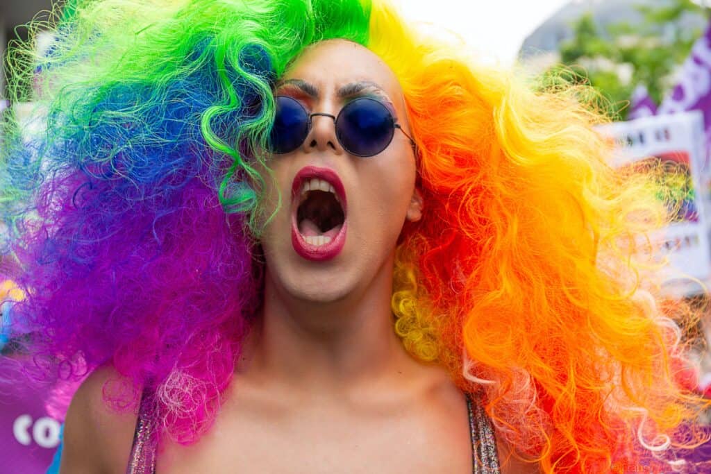 Individual with rainbow hair and sunglasses cheering