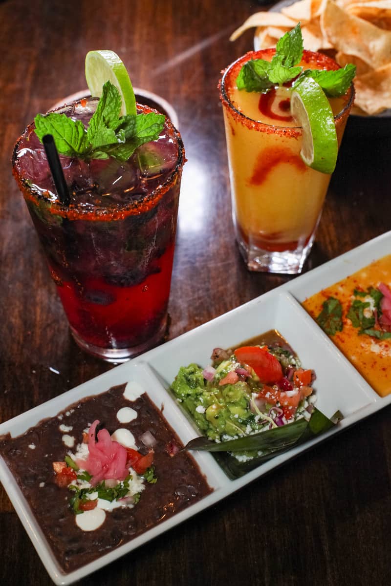 A blueberry mojito and mango margarita on a table behind a dish with three different dips in each of the three compartments. There is a black bean dip, guacamole dip, and cheese dip.