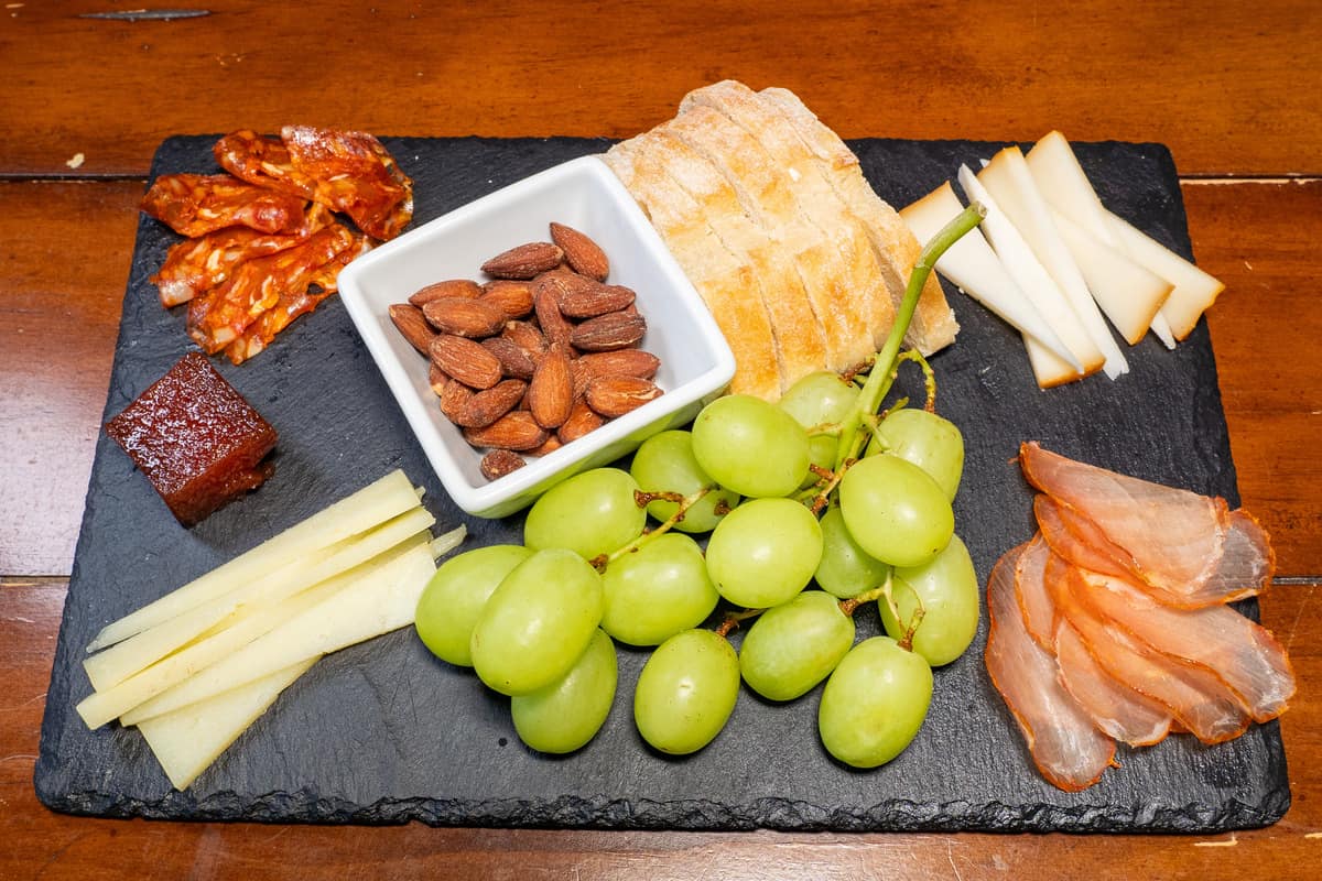 Charcuterie board on a black slate with thinly sliced meats, cheeses, green grapes, a cube shaped chunk of jam, a bowl of almonds, and pieces of sliced baguette.