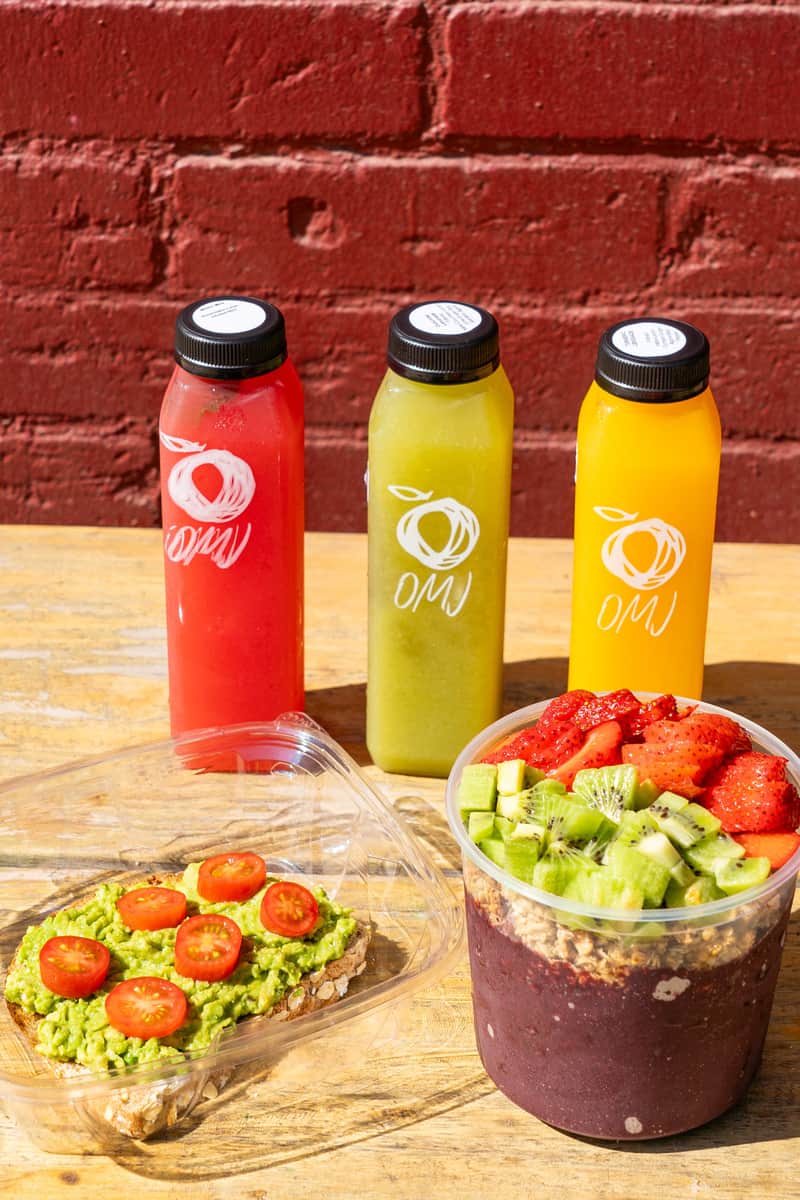 Small bottles of freshly squeezed juices behind a piece of avocado toast with sliced cherry tomatoes on it and a smoothie bowl. The juices are red, green, and yellow from left to right. The smoothie mixture in the bowl is purple, topped with granola, sliced kiwi, and strawberries.