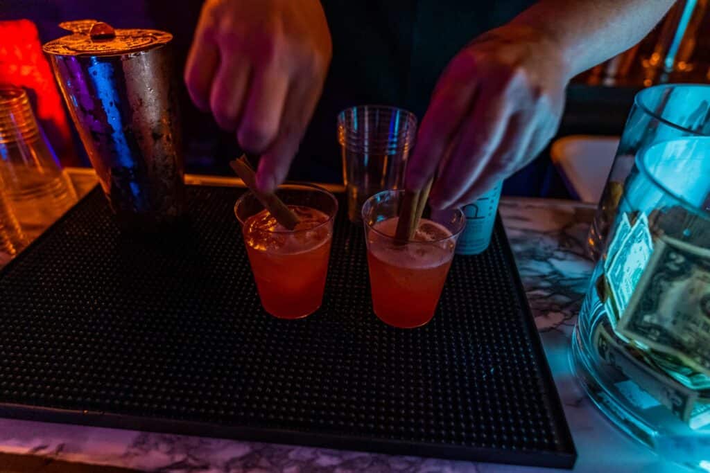 Two cocktails with cinnamon sticks in them being prepared by a bartender