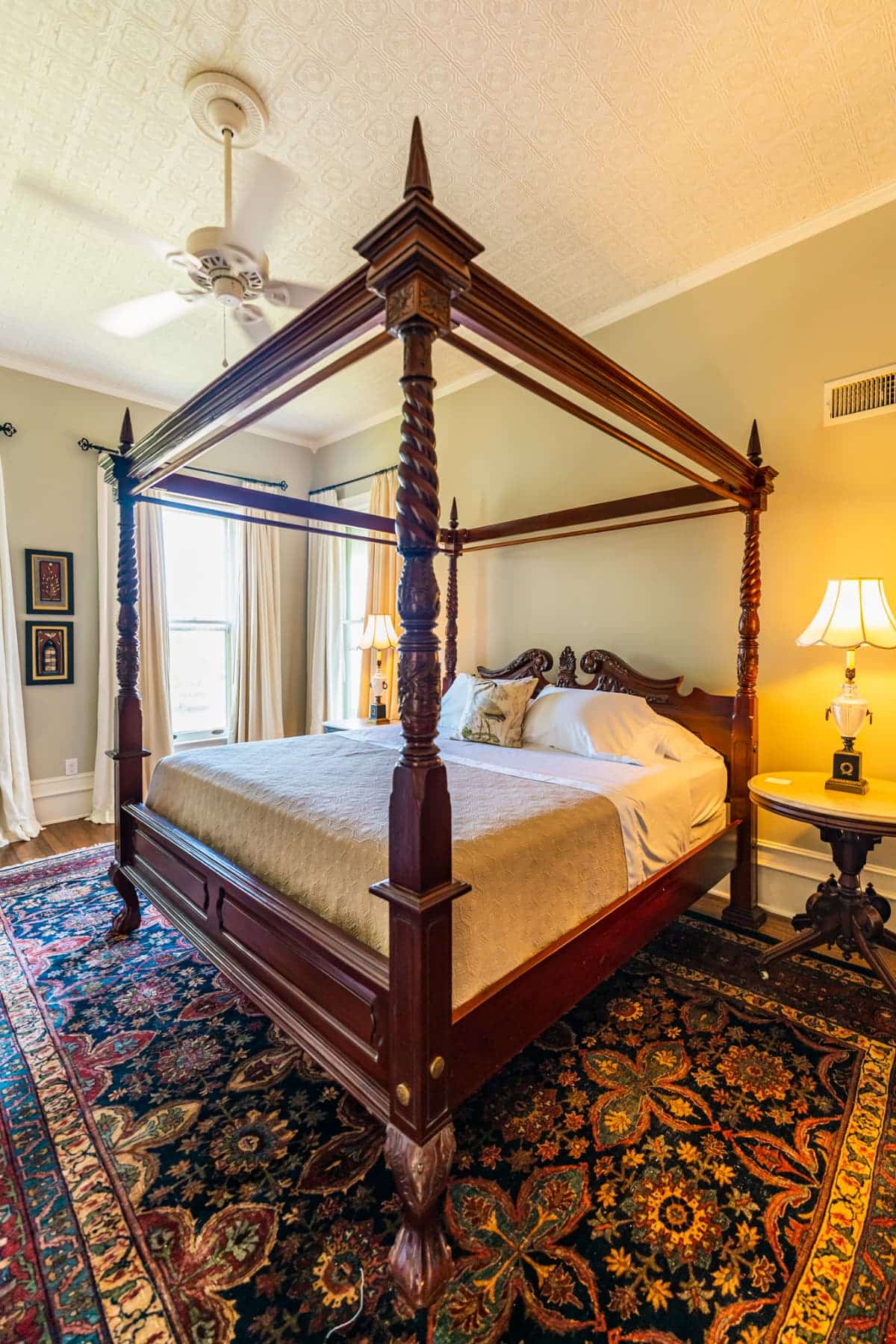 A wood canopy bed, a bedside table, and luxurious rug with the curtains open in the background