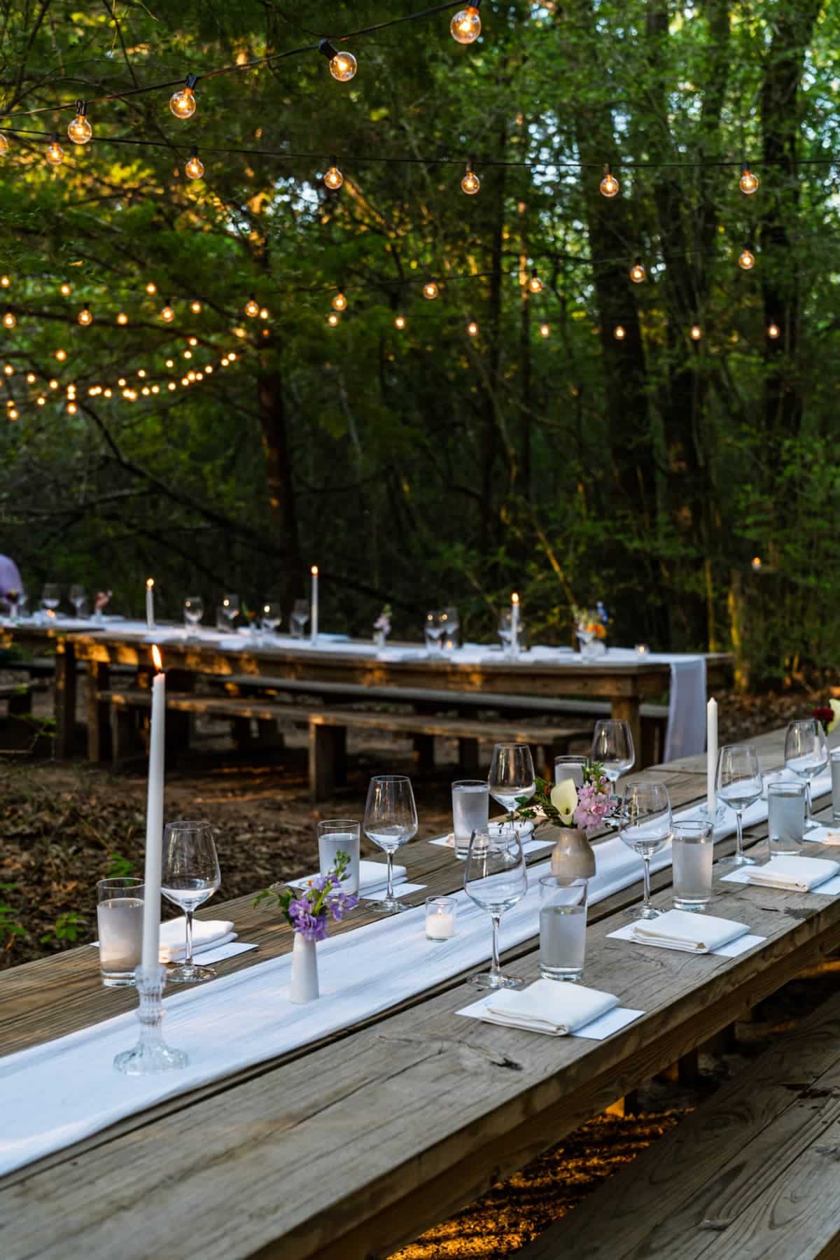 Two long wood tables in the middle of a forest and set with glasses, napkins, and lit candle sticks
