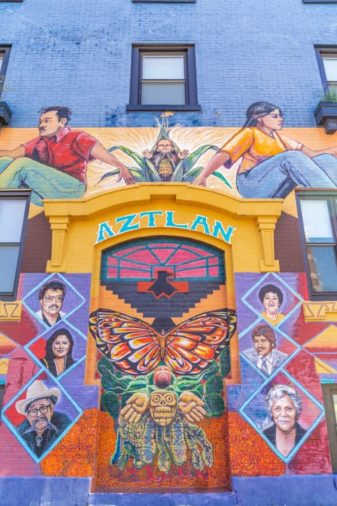 Bright mural on the side of a building of people's faces with "Aztlan" above it