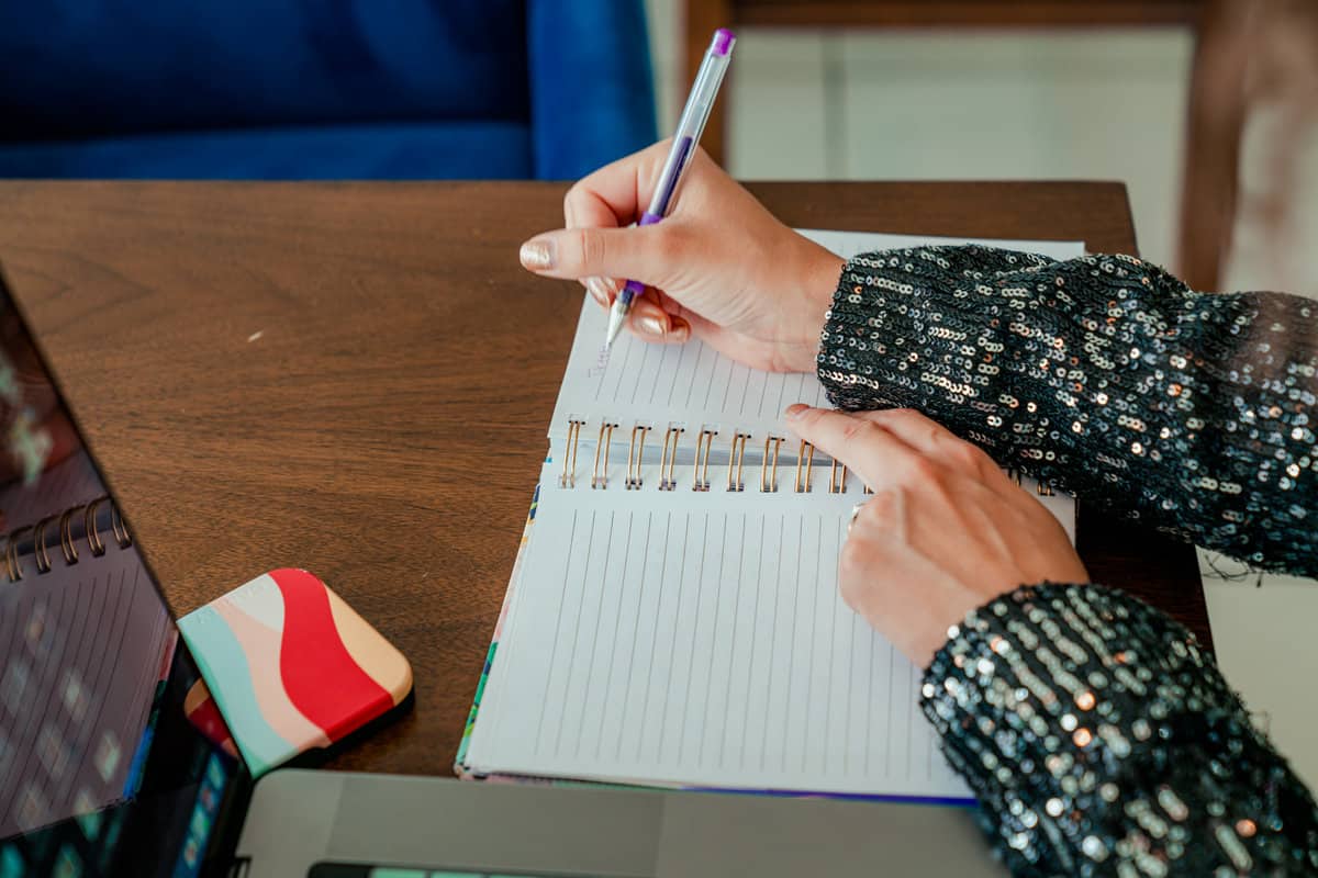 A woman sits at a table, writing in a notebook with a pen