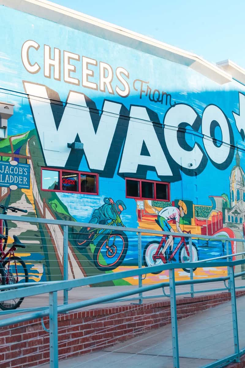 A mural on an exterior wall that says "Cheers from Waco" with depictions of people on bikes, the iconic courthouse, and other Waco landmarks