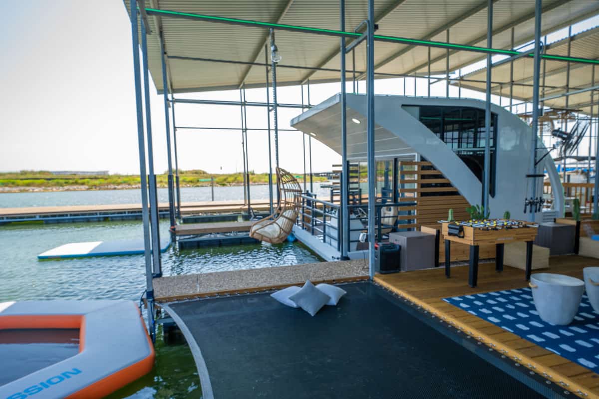 A floating dock with a modern lounge area, hanging chair, and spiral staircase under a metal roof.