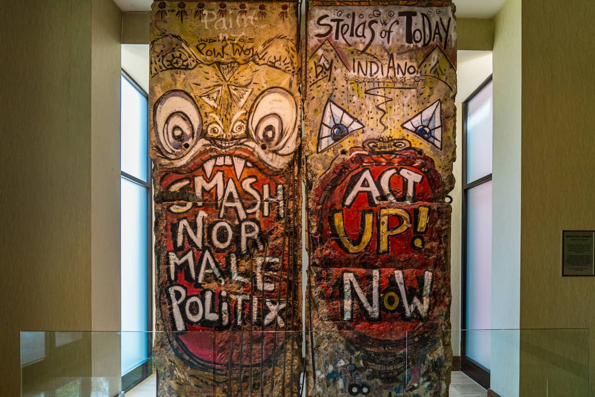 Graffiti-covered segment of the Berlin Wall displayed indoors with bold text and caricatures.