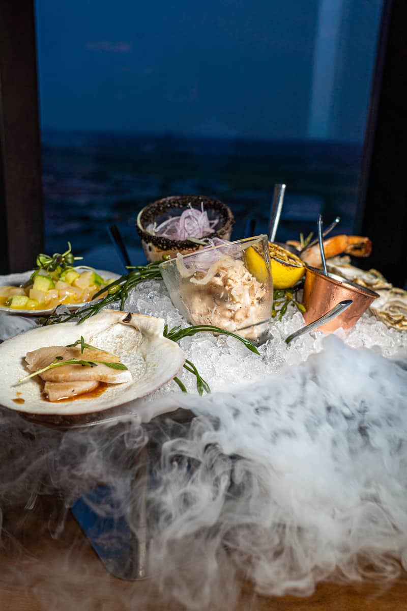 Assorted seafood dishes on ice with dramatic dry ice vapor, ocean backdrop.