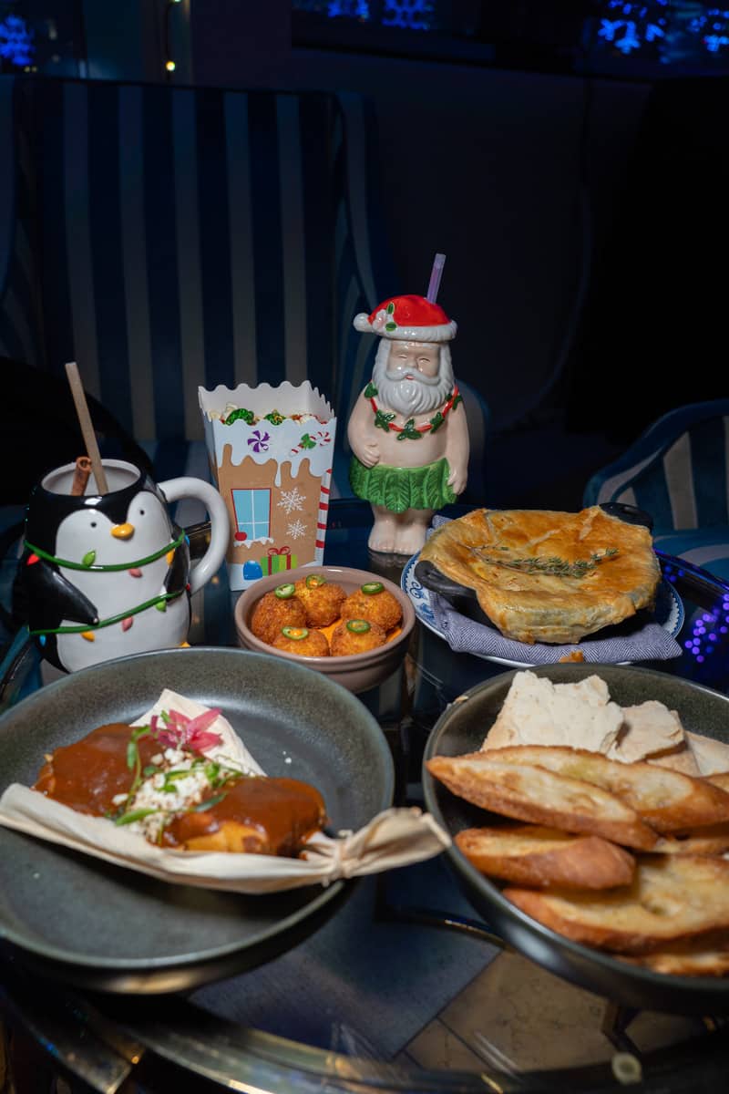Plates of food and three cocktails in novelty Christmas mugs on a table. One mug looks like Santa in a Hula skirt. The other two look like a penguin and a gingerbread house.