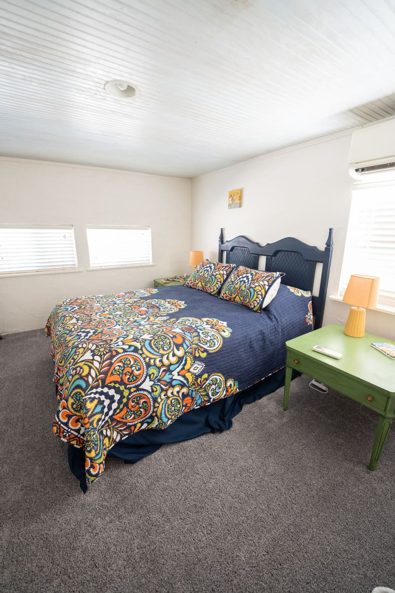 Navy blue bed spread with a colorful pattern near the foot of the bed. There is a green coffee table next to it. White walls surround it.