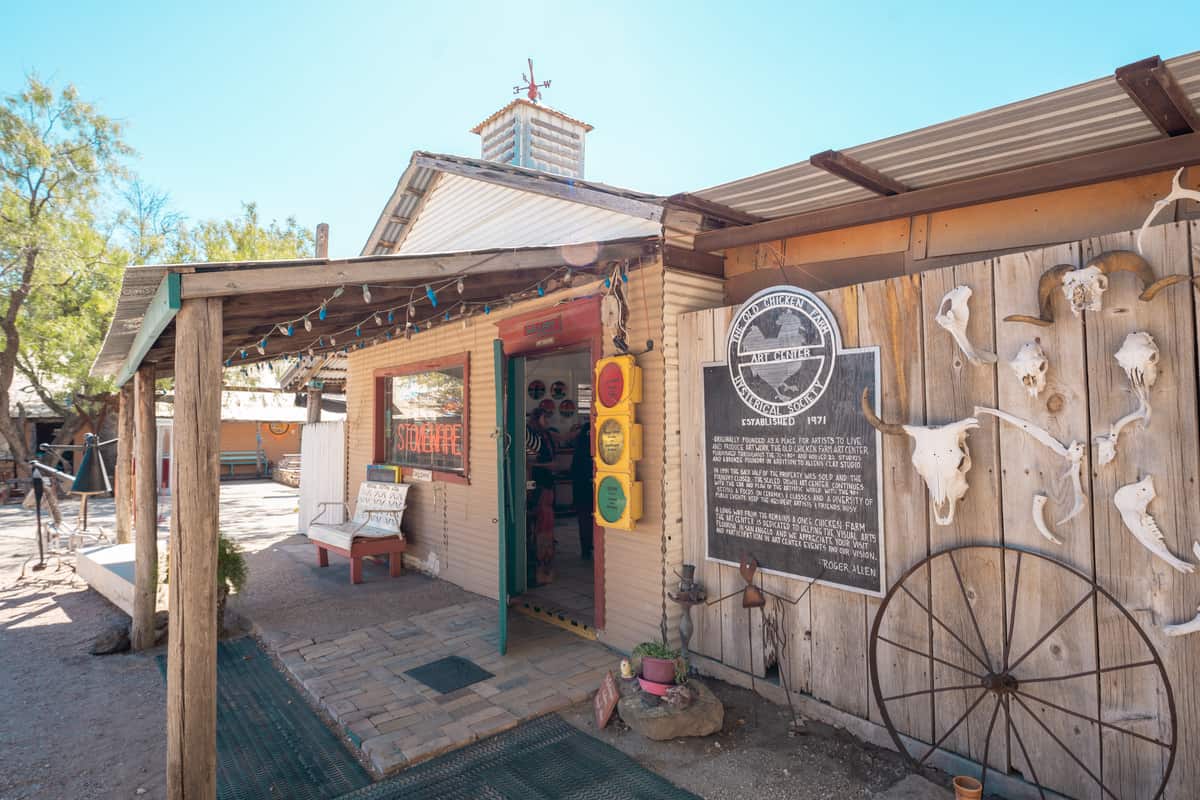 Exterior of the Old Chicken Farm Art Center. It's made of repurposed wood with lights hanging from the awning, animal skulls on the wall, and a sign describing the history of the property.