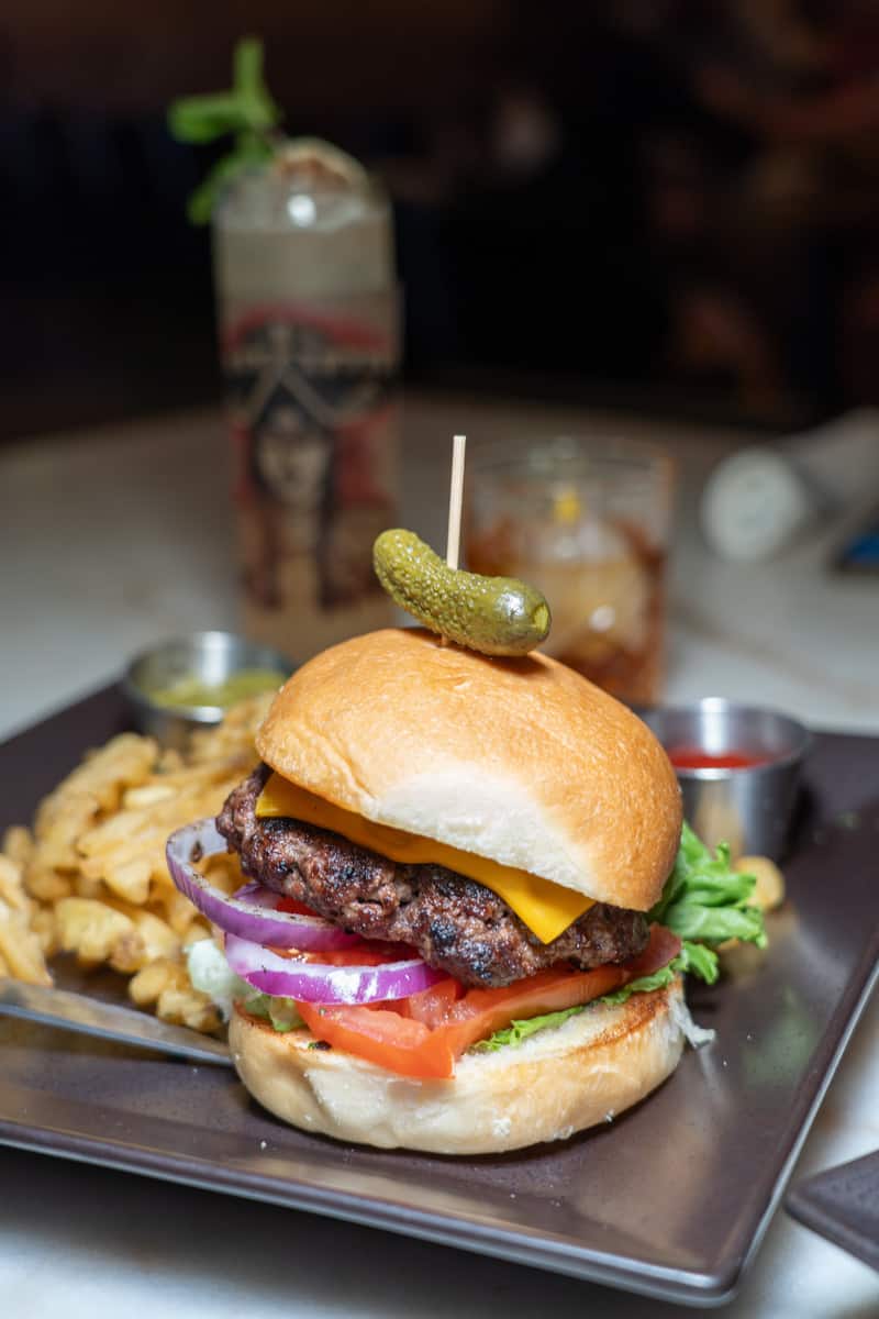 A burger on a plate with red onions, tomatoes, lettuce, and cheese on it. A Gherkin pickle is on top of it with a toothpick sticking out of it. Fries are on the plate beside it. There are two cocktail glasses in the background.