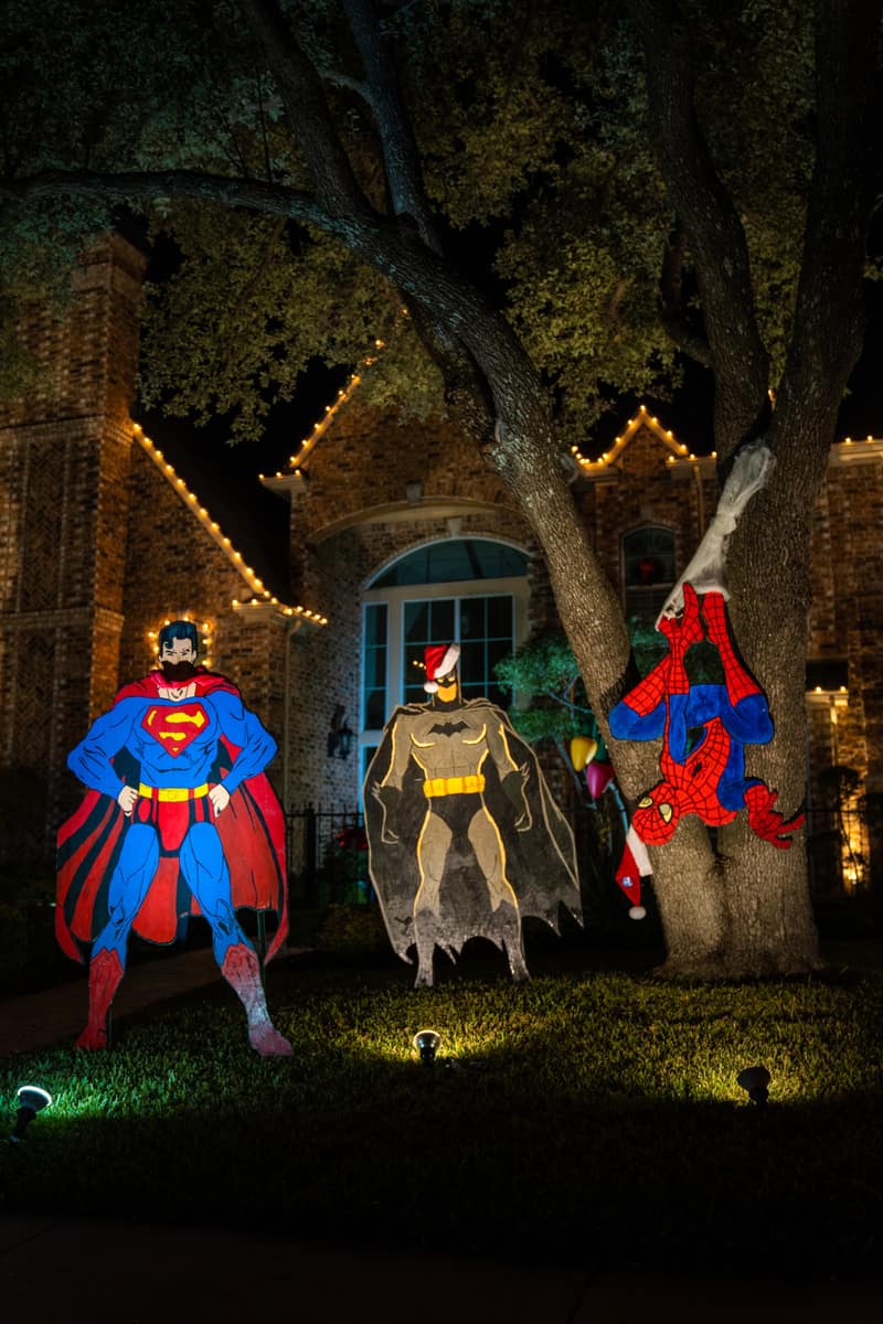 Cut-outs of Superman, Bat Man in a Santa hat, and Spiderman hanging from a tree on the lawn of a house illuminated by Christmas lights.