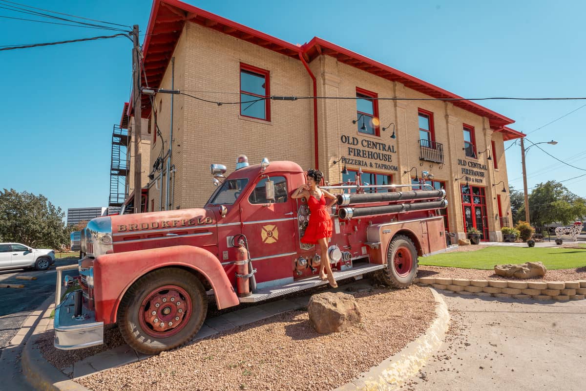 The front of the Old Central Firehouse Bed & Brew with an old fire engine in front of it. A woman in a red dress is hanging off the firetruck.