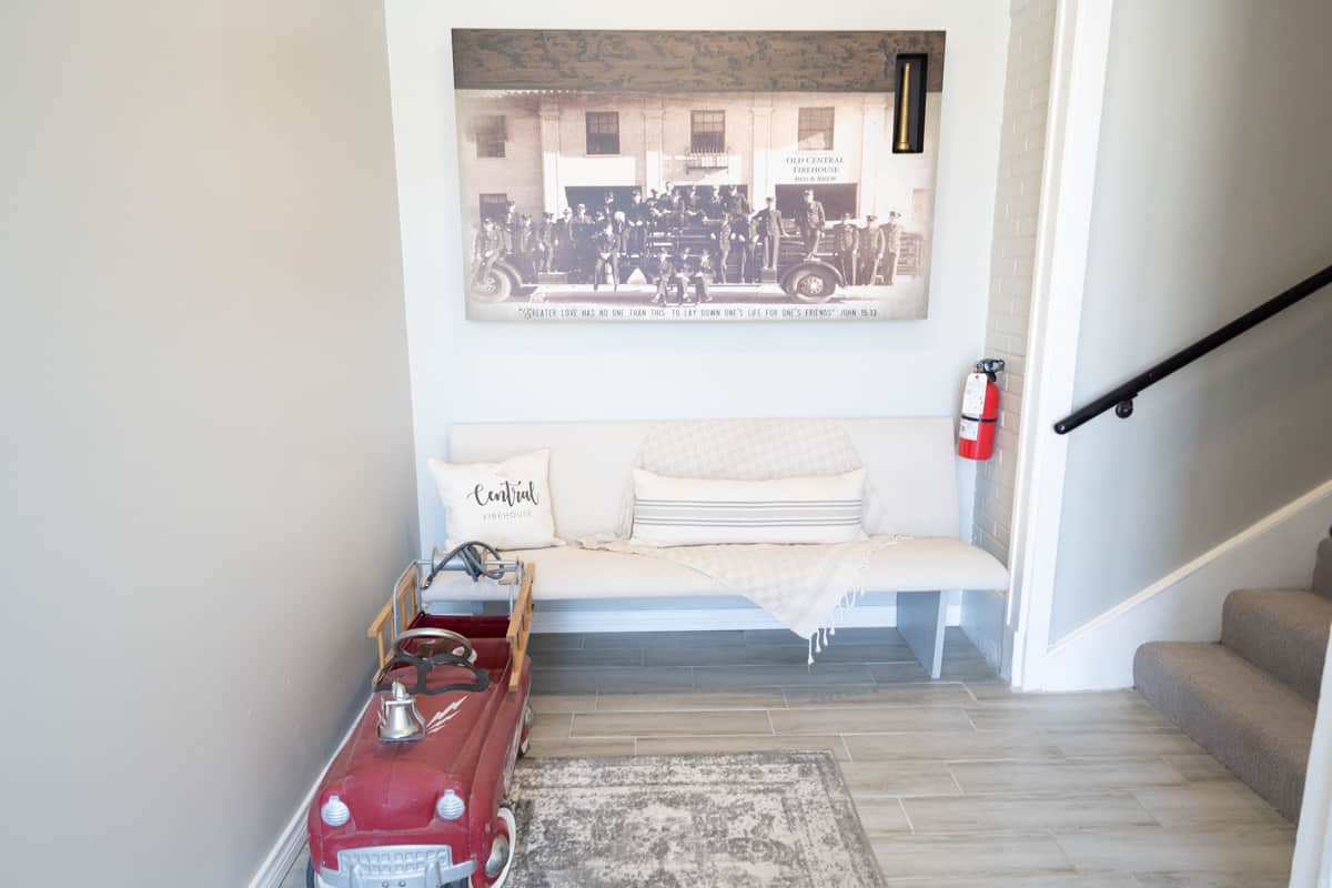 A white couch with its back to the wall and a black and white photo of the original firehouse above it. There is a staircase to the couch's left and a miniature antique fire engine on the floor in front of it.