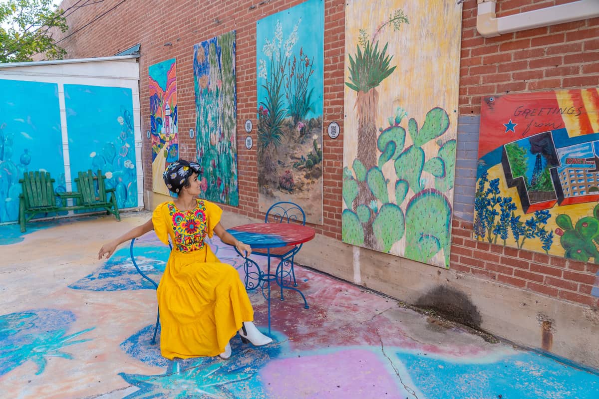 A woman sitting at a table surrounded by colorful art on the brick wall next to her and on the ground around her