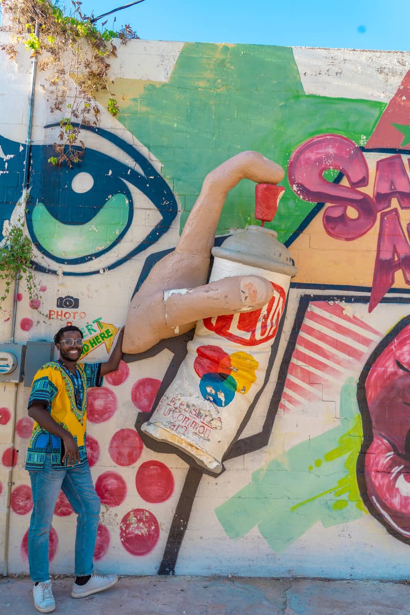 A man standing next to a colorful mural on a wall with a 3D hand and spray paint can coming out of the wall. The hand is spray painting the words "San Angelo" on the wall.