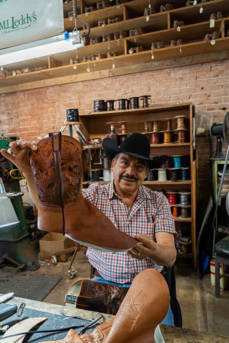 A man in a black cowboy hat holding up a single incomplete brown cowboy boot he is working on.