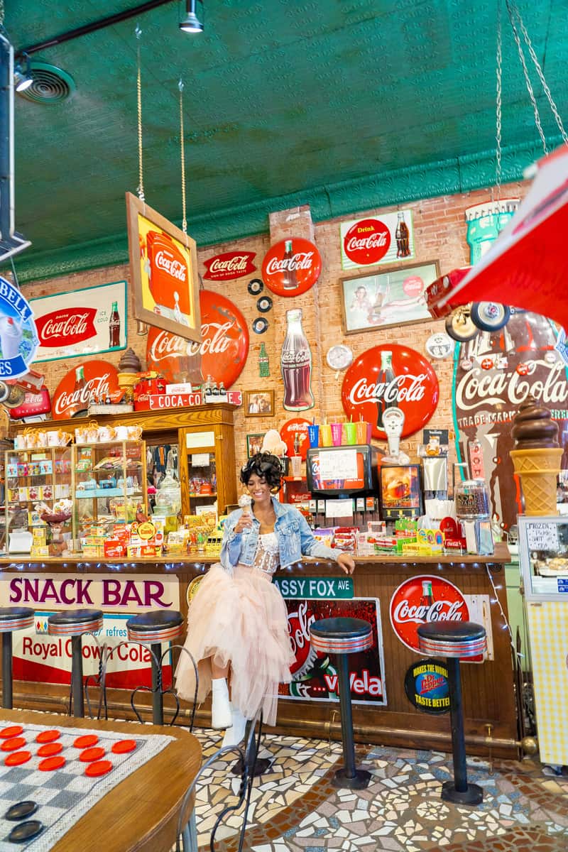 A woman sitting at an ice cream counter, holding an ice cream cone. There are lots of antique signage all around, including different Coca-Cola signs on the wall behind her.