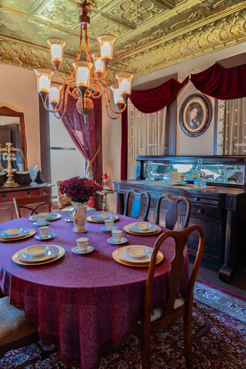 A round table covered in a maroon tablecloth surrounded by wooden chairs. There are 6 table settings, one in front of each chair. There is antique furniture on each wall with a multi-bulb chandelier hanging from the ceiling.