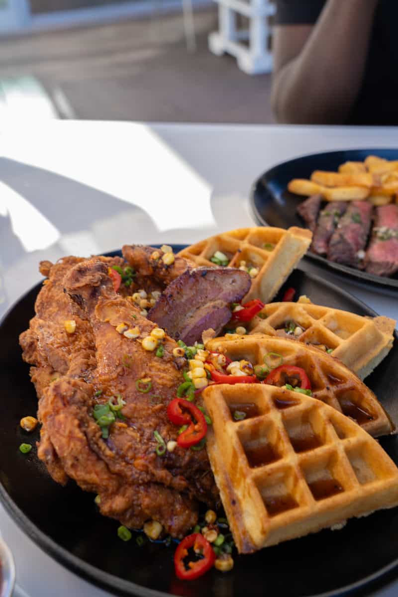 A plate of chicken and waffles with corn and roasted red peppers sprinkled on top