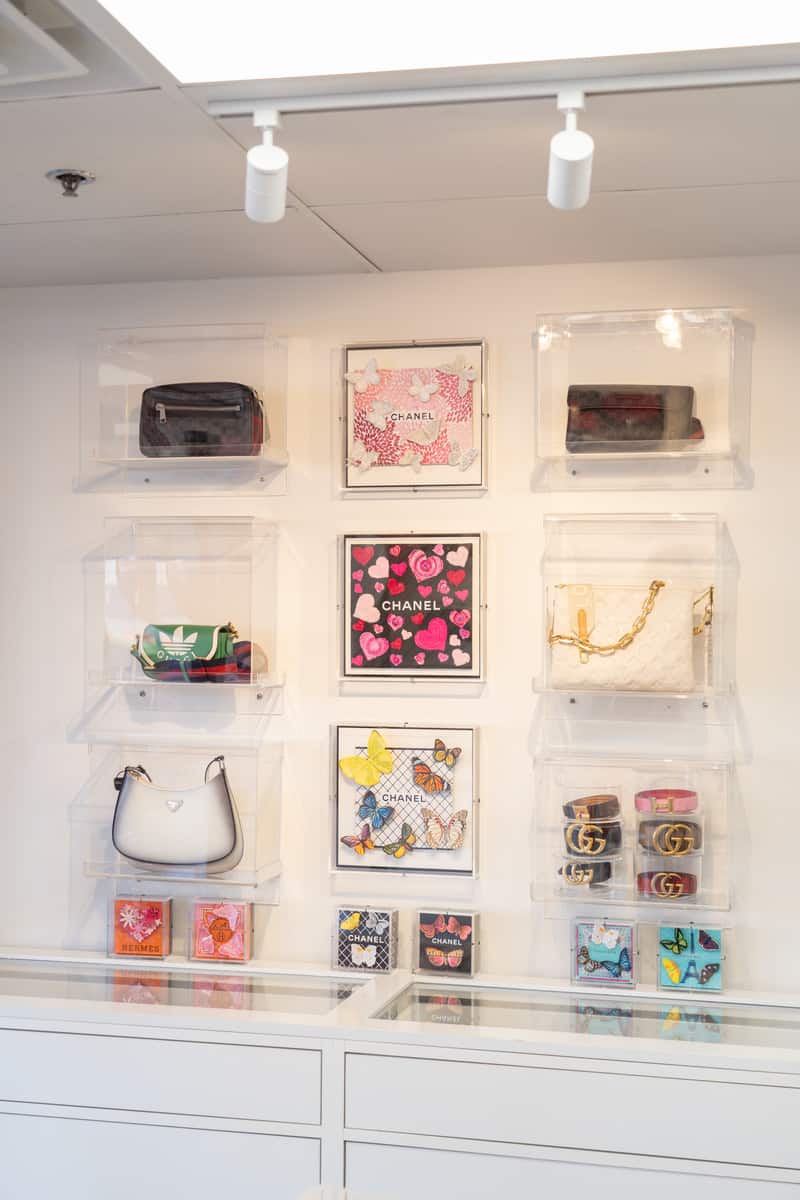 Designer purses and belts on display on the wall in clear boxes.