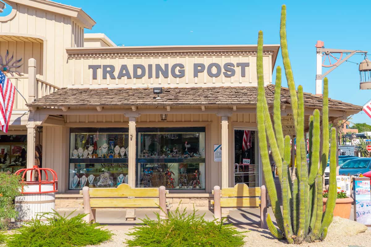 A historic looking storefront with a tall cactus and a sign that says "Trading Post" on it.
