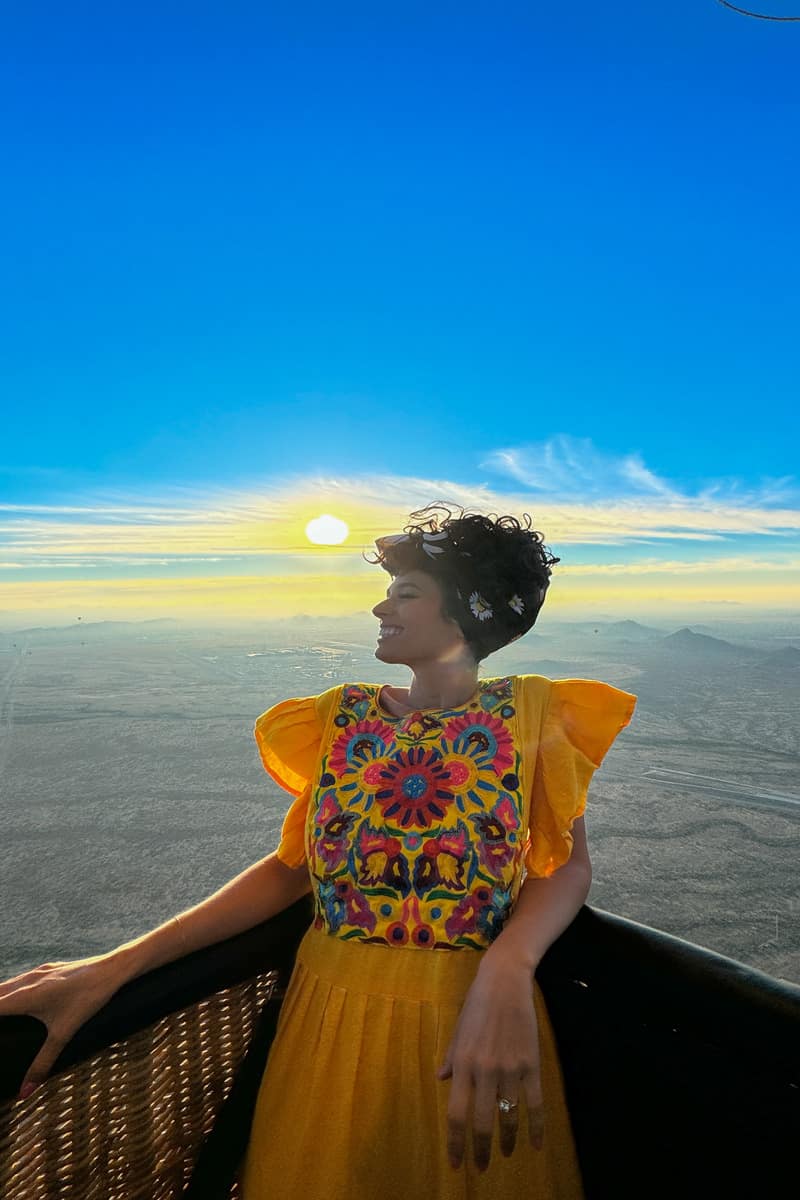A woman standing in a hot air balloon basket with the landscape and a sunrise behind her.