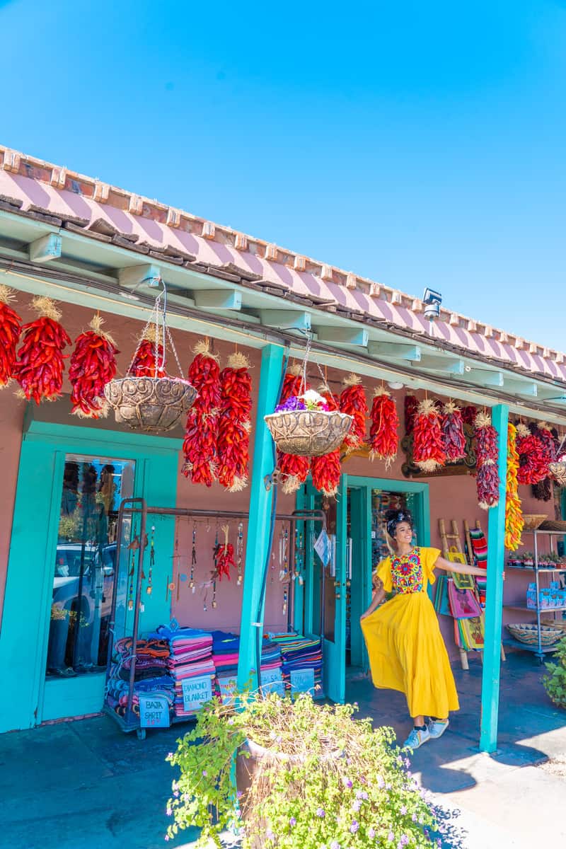 A woman standing on the porch of a colorful storefront with dried peppers hanging in bushels from the roof