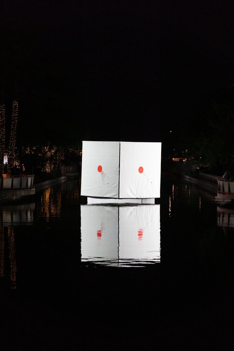 What looks like a white cube with a red circle in the center of each of its faces, reflecting in the water beneath it.