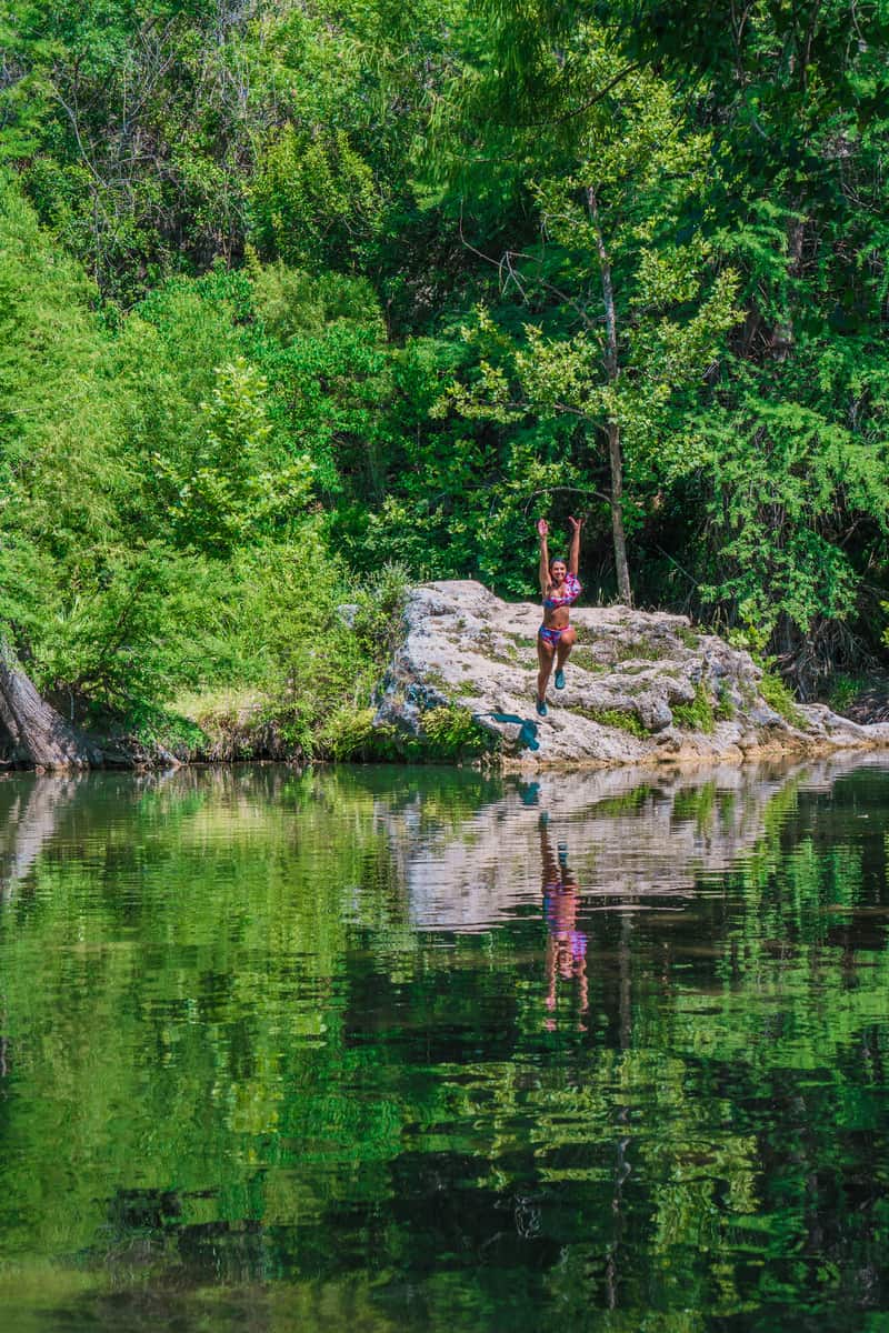 A woman in a bathing suit jumping into the water from a small boulder with green trees growing behind it