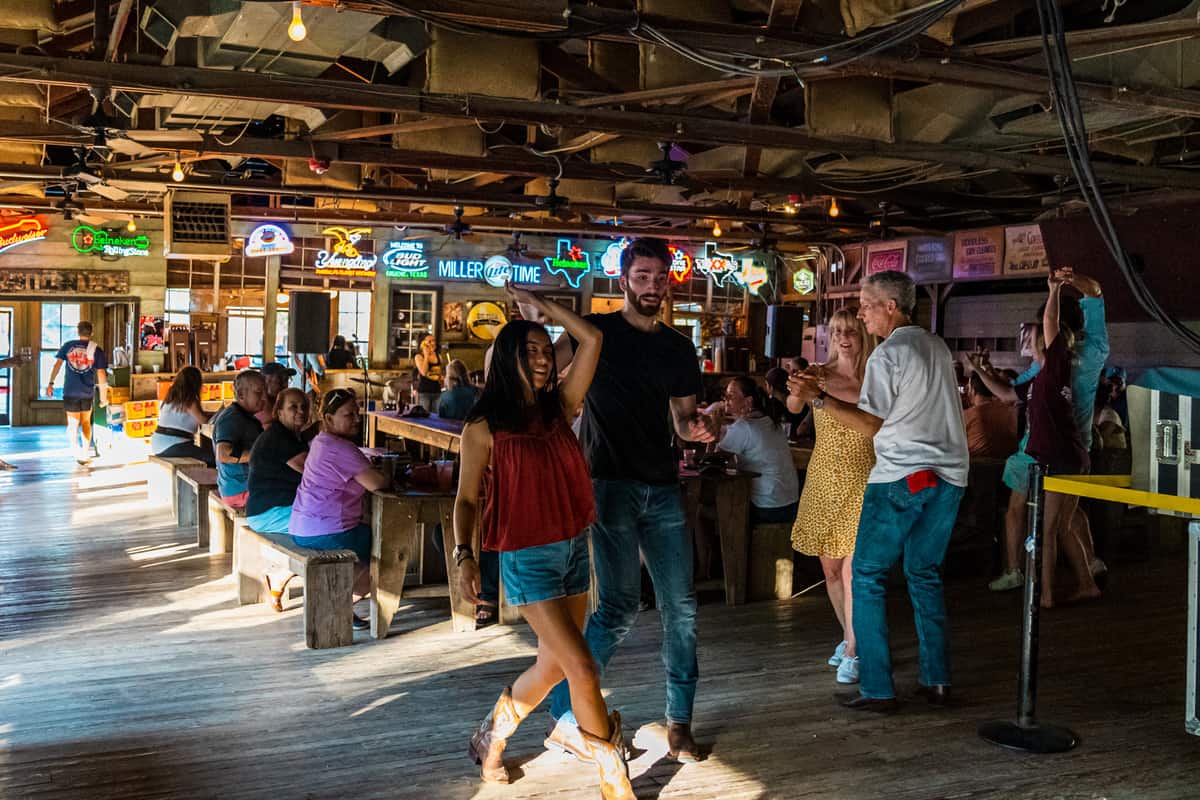 Couples dancing on a wooden dance floor with people sitting at long, wooden tables behind them