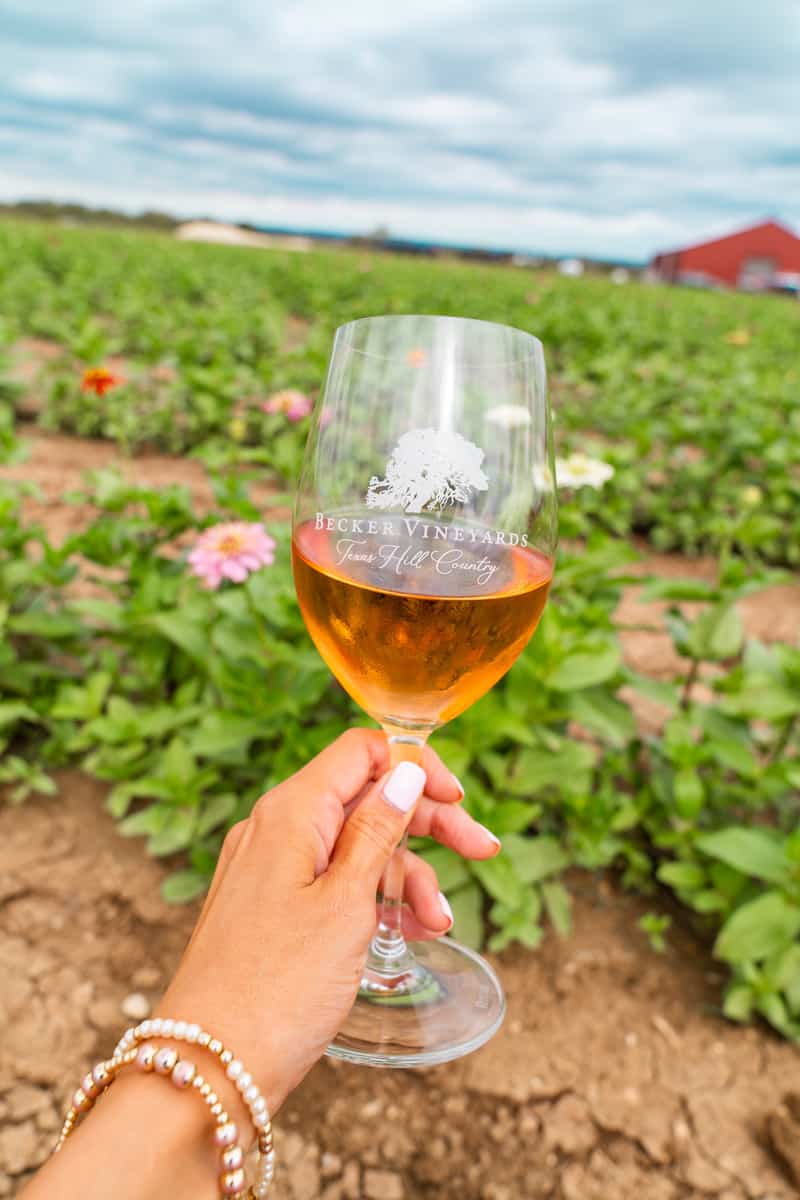 A wine glass held by a womans hand with a backdrop of a flewer garden.