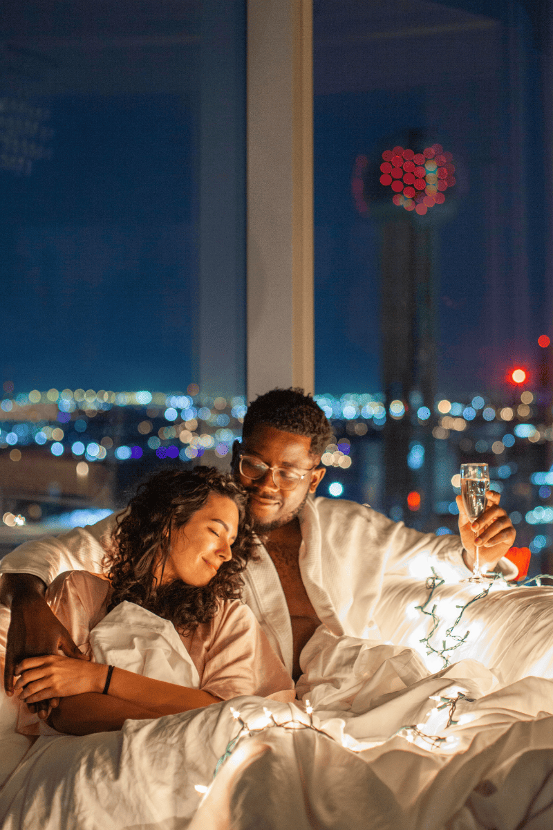 A couple in bed with lights on, enjoying the city view from their window.


