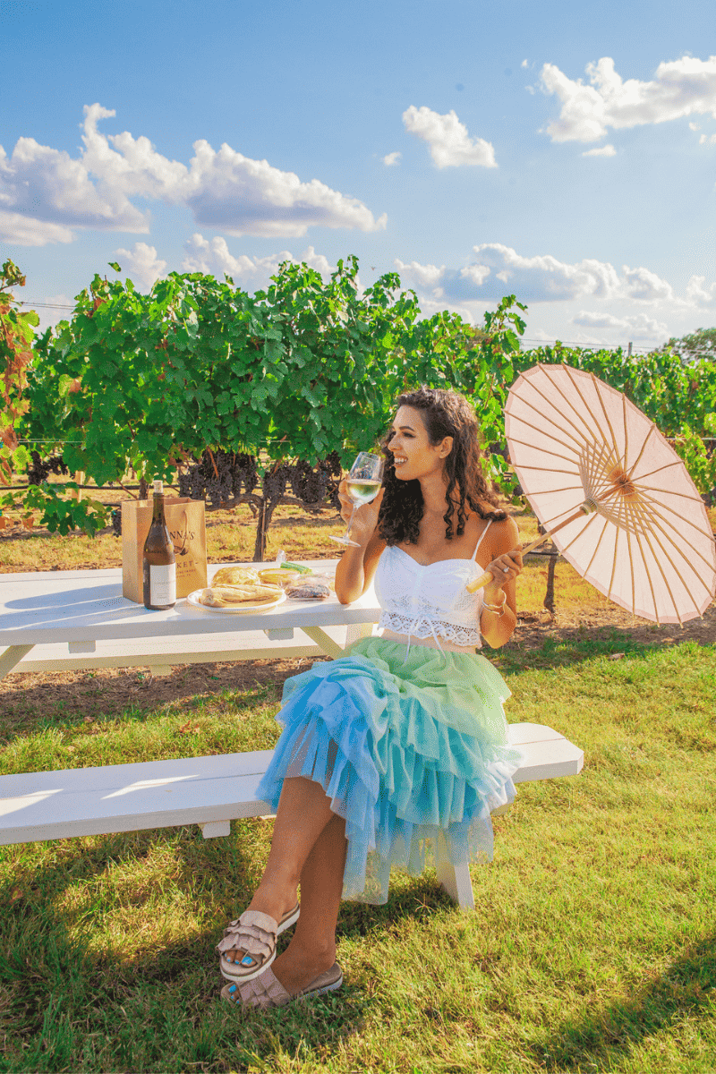 A woman in blue skirt and white top holding an umbrella enjoying a glass of wine with a vineyard on the background