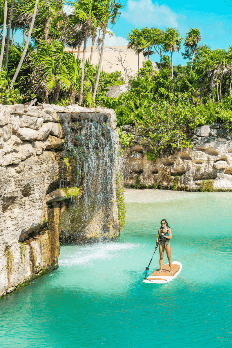 A woman paddle boarding near a waterfall, gracefully gliding on the water's surface with the scenic cascade in the background.