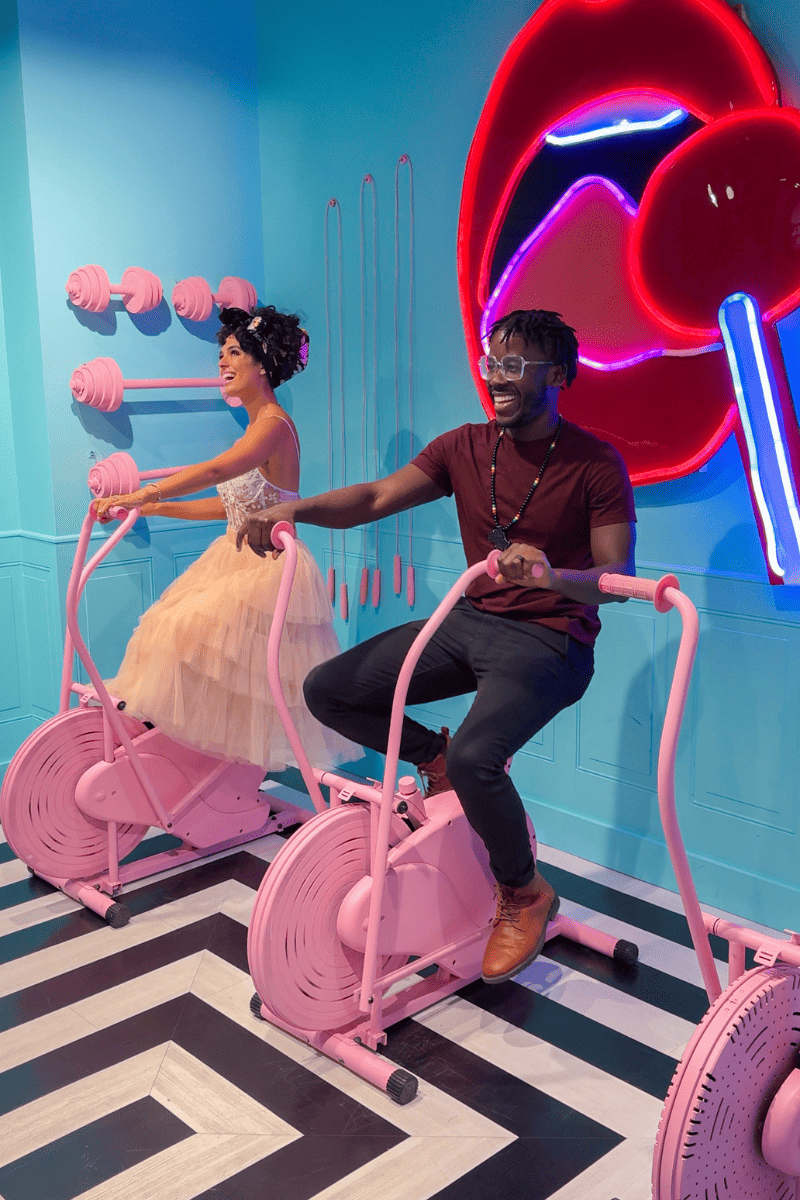 Couple riding a pair on pink exercise bikes in neon-lit room