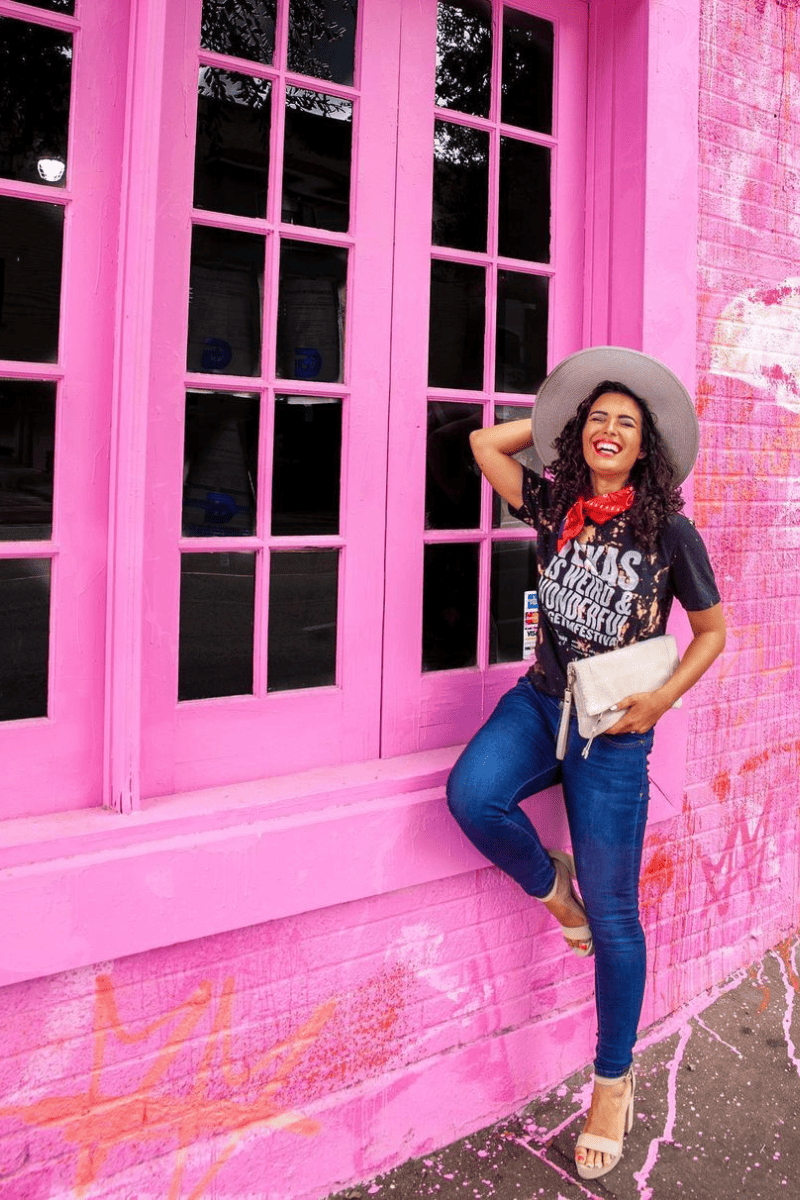 A woman in jeans and black top and a handy bag happily making a pose on an pink building