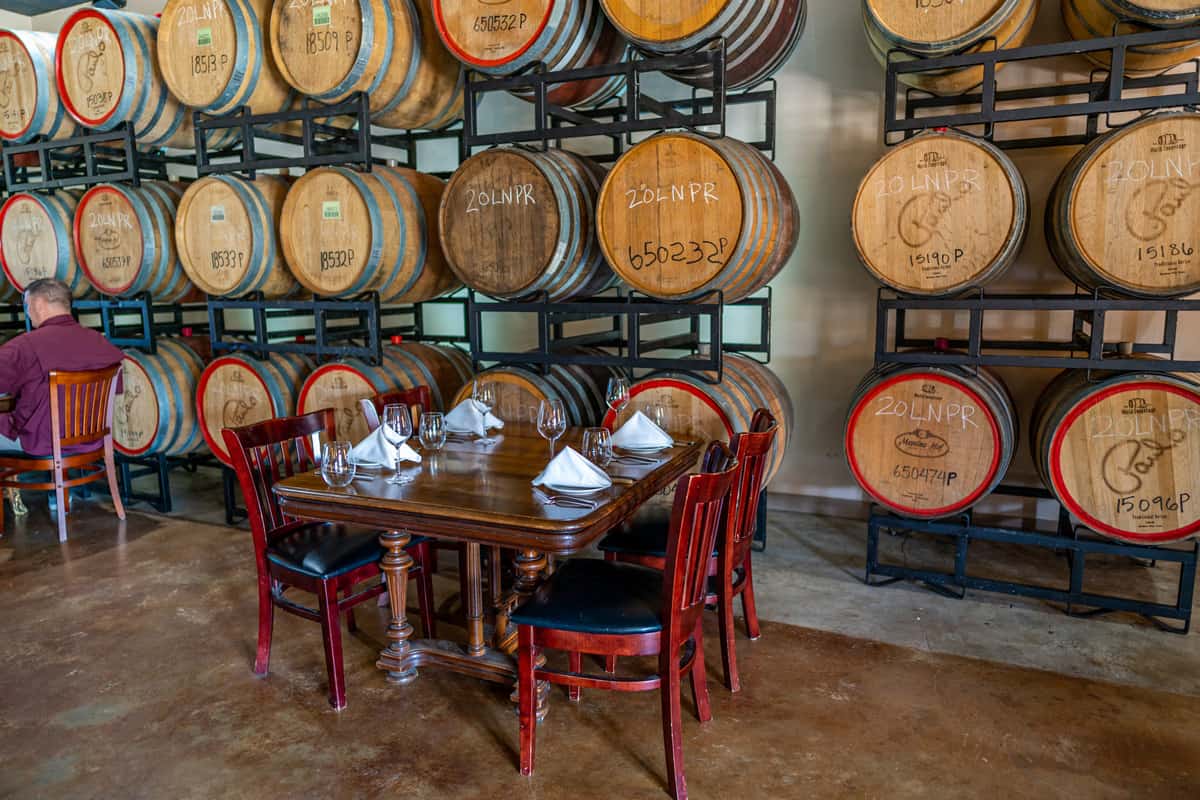 A table with four chairs in front of a wine barrel filled room.