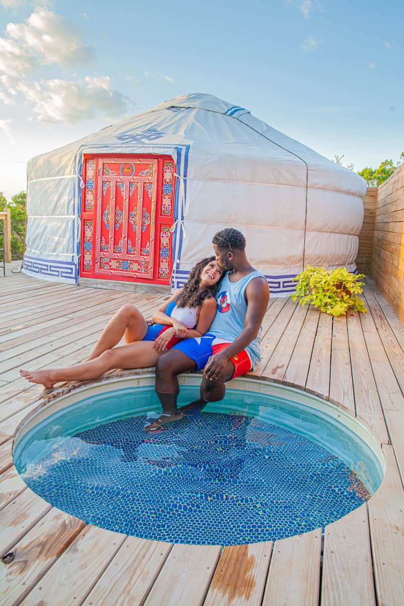 A couple relaxes together in a small circular pool beside a traditional yurt