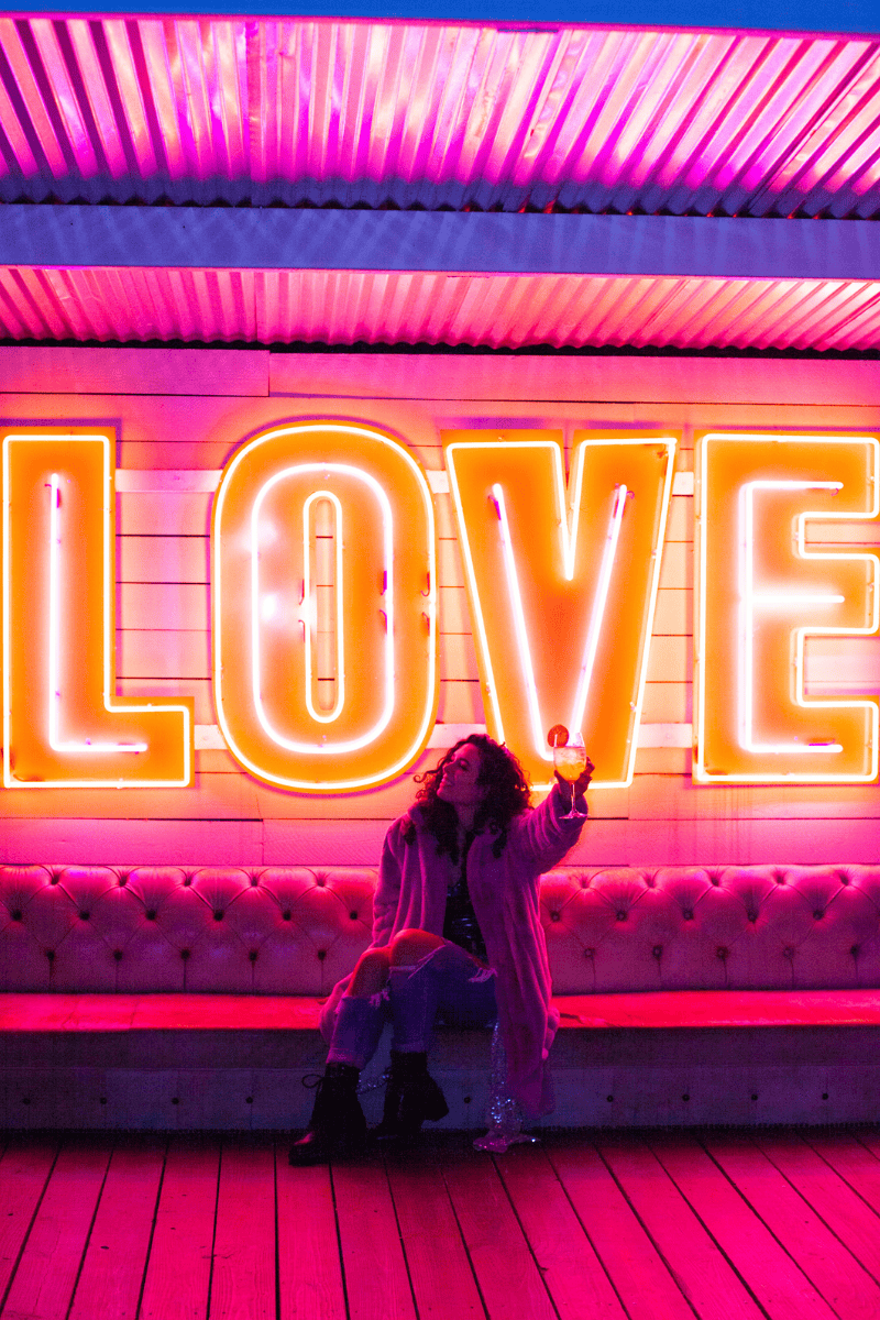 A woman sitting on a bench in front of a neon love sign